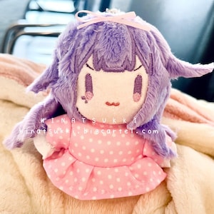 May include: A plush doll with a lavender-colored head and hair, wearing a pink polka-dot dress. The doll has embroidered facial features and a small pink bow. The text "INATSUKKI" and "inatsukki.bigcartel.com" are visible.