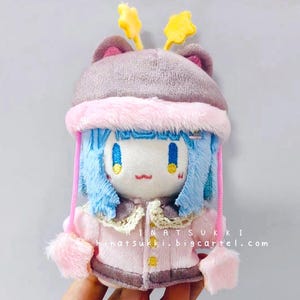 May include: A plush doll with blue hair, wearing a pink coat and a gray hat with yellow star accents. The doll has a white face with blue eyes and a small red mouth. The text "HINATSUKKI hinatsukki.bigcartel.com" is visible.