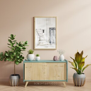 May include: A light wood cabinet with two doors and a light blue top. The cabinet is sitting on a light wood floor. There are three plants in pots on top of the cabinet. A framed watercolor painting of a white building with a door and stairs is hanging on the wall behind the cabinet.