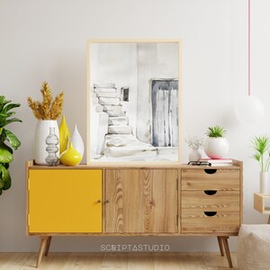 May include: A watercolor painting of a white building with a doorway and steps, framed in a light wood frame, hanging above a wooden cabinet with a yellow door and three drawers.