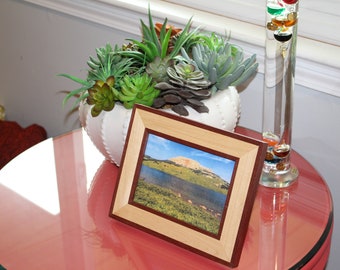 May include: A square framed picture with a landscape scene of a mountain and lake. The frame is light wood with a dark wood border. A white ceramic pot with succulents sits behind the frame. A glass thermometer is to the right.