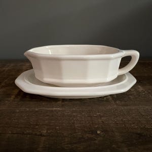 May include: A white, octagonal gravy boat with a handle, resting on a matching saucer. The set is made of ceramic and sits on a dark wooden surface. The gravy boat has a classic, elegant design.