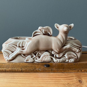 May include: A ceramic planter in the shape of a deer leaping over foliage. The planter is a light beige color with brown accents, and has a rectangular base with two openings for plants. The deer is in mid-leap, adding a dynamic element to the design.