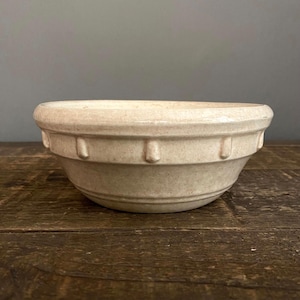 May include: A beige ceramic mixing bowl with a wide rim and a series of vertical indentations around the middle. The bowl has a rounded shape and sits on a wooden surface. The bowl is a neutral color.