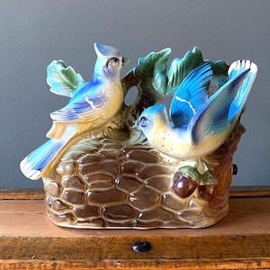 May include: A ceramic bird figurine featuring two blue and yellow birds perched on a brown, textured base with green leaves. The birds have detailed feathers and are set against a neutral background. A decorative piece.
