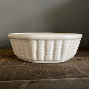 May include: A round, off-white ceramic bowl featuring a raised, textured pattern on the exterior that mimics the appearance of woven basketry. The bowl has a smooth, thick rim and a consistent, glossy finish across its entire outer surface.