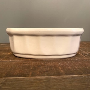 May include: A white ceramic baking dish with a smooth, glossy finish and a rounded, fluted exterior design. The dish features a thick, reinforced rim and a flat base, presenting a clean, solid color appearance suitable for various kitchen tasks.