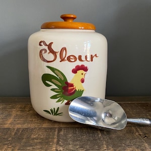 May include: Cream-colored ceramic flour canister with a wooden lid and a metal scoop. The canister features a rooster illustration and the word "Flour" in brown script. The scoop is silver and rests on a wooden surface.