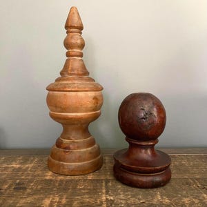 May include: A tall, light brown wooden finial with a pointed top and a rounded base, positioned next to a shorter, dark brown wooden sphere mounted on a circular pedestal base. Both objects feature smooth, turned wood construction and visible grain.