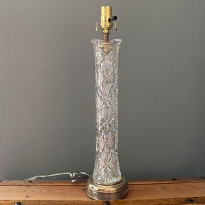 May include: A clear, cut-glass table lamp with a brass base and lamp holder. The lamp has a long, cylindrical body with a diamond-cut pattern. The lamp is approximately 20 inches tall.