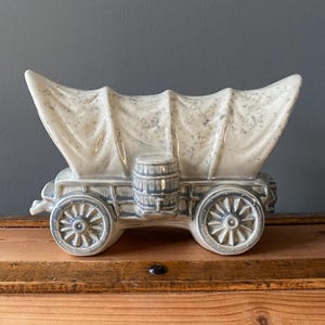 May include: A ceramic covered wagon planter in shades of gray and white. The wagon features spoked wheels, a barrel, and a canvas top. The decorative piece is designed to hold plants or other small items.