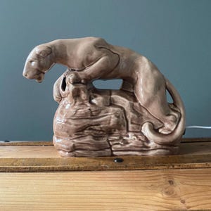 May include: A ceramic sculpture of a cougar perched atop a textured rock formation. The cougar is a light brown color, with detailed features. The rock formation is a darker brown, adding contrast. The sculpture is a decorative item.