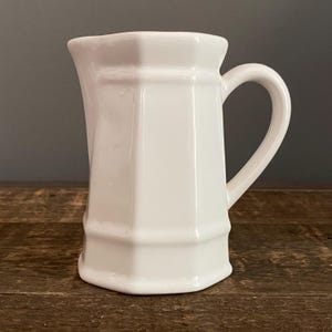May include: A white ceramic pitcher with a geometric design. The pitcher has a handle and a spout. The pitcher is sitting on a wooden surface.
