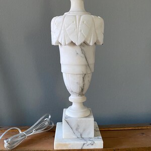 May include: A white marble table lamp with grey veining, featuring a carved leaf-patterned upper section, a tapered body, and a square base. The lamp includes a clear electrical cord with a plug, resting on a flat surface.