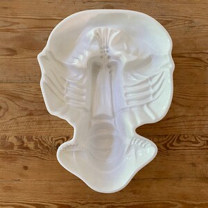 May include: A white ceramic serving dish with a face-like design. The dish has a long, vertical central section resembling a nose, flanked by wing-like shapes. The overall form is symmetrical and rests on a wooden surface.