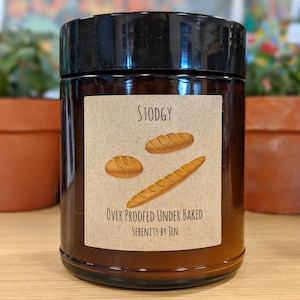 以下が含まれることがあります： 「Stodgy」の文字とパンのイラストが描かれたラベルが付いた、黒い蓋の茶色のガラス瓶。ラベルには「Over Proofed Under Baked」と「Serenity by Jen」の文字も。瓶は木製の表面に置かれています。