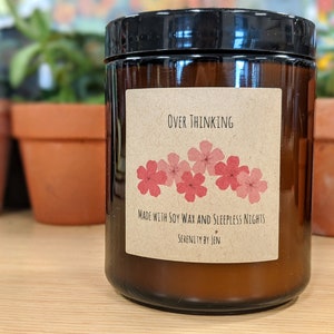 May include: A brown glass candle jar with a black lid. The label reads "Over Thinking" with pink flower illustrations. The text below says "Made with Soy Wax and Sleepless Nights" and "Serenity by Jen".