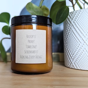 May include: A brown glass jar candle with a black lid and a beige label. The label has the words "RECEIPTS! PROOF! TIMELINE! SCREENSHOTS! FUCKING EVERYTHING!" printed in black. The candle is on a wooden surface.
