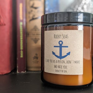 Op de afbeelding: Een bruine glazen potkaars met een zwart deksel. Het etiket leest "Rocky Seas" met een blauwe ankerafbeelding en de tekst "I like you as a person, don't make me hate you." De kaars is omringd door boeken.