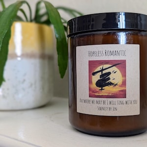 以下が含まれることがあります： 黒い蓋が付いた茶色のガラス瓶キャンドル。ラベルには「Hopeless Romantic」と書かれており、夕焼けを背景にしたシルエットのイラストが描かれています。「Anywhere we may be I will sing with you」と「Serenity by Jen」の文字も表示されています。