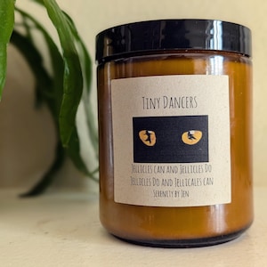 Puede incluir: Una vela en tarro de cristal con tapa negra y una etiqueta que dice "Tiny Dancers". La etiqueta presenta un rectángulo negro con dos ilustraciones de ojos de gato amarillos. El texto "Jellicles Can and Jellicles Do" también está en la etiqueta.