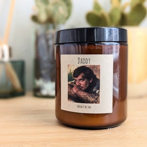 Può includere: Un barattolo di candela in vetro marrone con coperchio nero. L'etichetta recita "DADDY" sopra una foto vintage di un uomo. Il testo "SERENITY BY JEN" è sotto. La candela è su una superficie di legno.