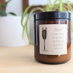 May include: A brown glass candle jar with a black lid and a label that reads "DÉCLASSÉ I'M NOT SAYING THIS AS A THREAT, I'M SAYING THIS AS A PROMISE." The label also features a champagne glass illustration. The candle is on a light wood surface.