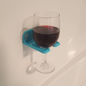 May include: A blue plastic wine glass holder attached to a white tiled wall with a glass of red wine inside.