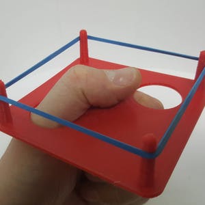 May include: A red plastic boxing ring with blue rubber bands for ropes. The ring is designed to fit around a finger.
