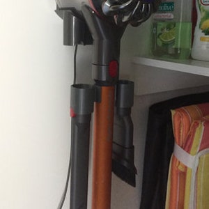 Könnte beinhalten: Ein grauer und orangefarbener Akku-Staubsauger ist an einer Wand mit einer Ladestation montiert. Der Staubsauger hat einen blauen und silbernen Kopf und einen roten Knopf. Der Staubsauger befindet sich in einer Küche oder einem Badezimmer.