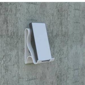 May include: White plastic phone stand mounted on a textured gray wall. The stand is designed to hold a phone in a vertical position.