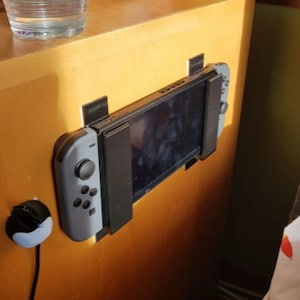 Puede incluir: Una consola Nintendo Switch gris con un mando negro está montada en una superficie de madera clara. La consola está fijada a la superficie con tiras adhesivas negras.