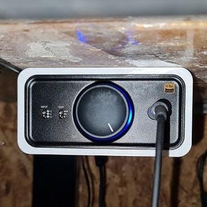 Puede incluir: Una interfaz de audio en blanco y negro con una gran perilla central y un anillo iluminado azul. El dispositivo está montado debajo de una superficie de madera y tiene un cable de alimentación enchufado. El dispositivo tiene el logotipo Hi-Res Audio.