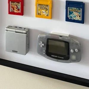 May include: A framed display featuring a collection of Game Boy Advance cartridges and two handheld consoles. The cartridges are in various colors, including red, green, blue, and yellow. The consoles are silver and gray.