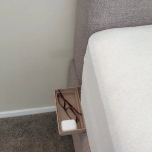 May include: A white bed with a gray headboard and a small beige shelf attached to the side. The shelf has a pair of glasses and a white rectangular object on it.