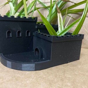 以下が含まれることがあります： アーチ型の小さな入り口と3つの窓が付いた、黒色の3Dプリントされた城の形をしたプランター。プランターには緑色の植物が植えられています。