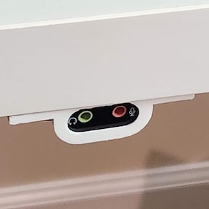 May include: A white, rectangular panel with a black audio port panel attached. The audio port panel has a green headphone icon and a red microphone icon. The panel is mounted on a beige surface.