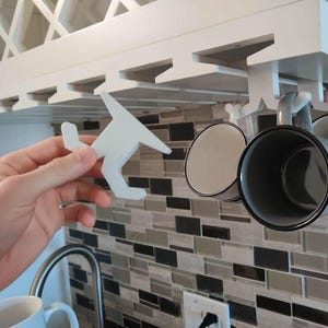 May include: A white wooden airplane-shaped mug holder is held near a white cabinet with a built-in mug rack. Three mugs hang from the rack. The mugs are gray with black rims and white interiors. The background is a mosaic tile backsplash.