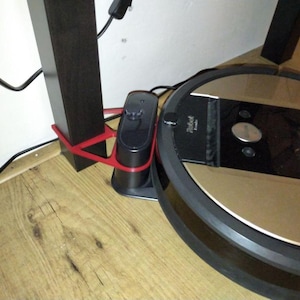 May include: A robotic vacuum cleaner with a black and gold design is docked next to a charging station. The charging station is black and red, and is attached to a dark brown support beam. Electrical cords are visible.
