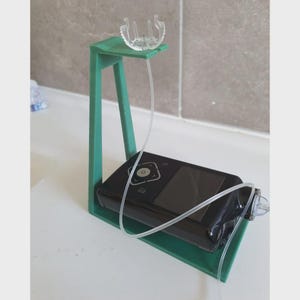 May include: A black medical device with a screen and buttons is held by a green stand. A clear tube connects the device to a clear plastic piece at the top of the stand. The stand is on a white surface.