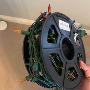 May include: A spool of multi-colored Christmas lights with red, blue, and yellow bulbs. The lights are wrapped around a black plastic reel. The reel is held by a person.
