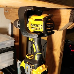 Può includere: Un trapano avvitatore a batteria DeWalt 20V MAX XR nero e giallo è tenuto in un supporto nero attaccato a una trave di legno. Il trapano ha un'impugnatura nera e un corpo giallo con il logo DeWalt. È un elettroutensile.