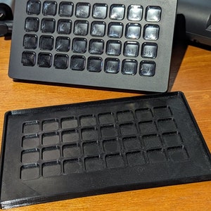 May include: Two black plastic trays with a grid of square indentations. The trays are designed to hold small objects, such as buttons or beads.
