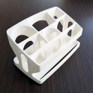 May include: White plastic organizer with multiple compartments, designed for storing small items. The organizer has a tiered design with a lower shelf and a top section with several open slots.