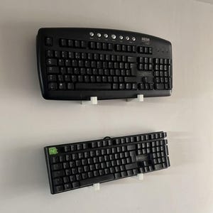 May include: Two black computer keyboards mounted on a white wall. The top keyboard is a full-size model with the brand name "MESH" visible. The bottom keyboard is a smaller, mechanical keyboard with the brand name "ROCCAT".