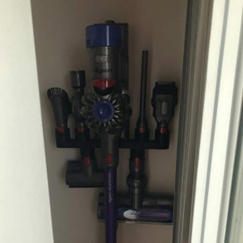 May include: A black wall-mounted storage rack for a cordless vacuum cleaner and its attachments. The rack has six slots for holding the vacuum cleaner, a crevice tool, a dusting brush, a combination tool, and two other attachments. The vacuum cleaner is a dark gray and purple color with a blue and red dustbin.