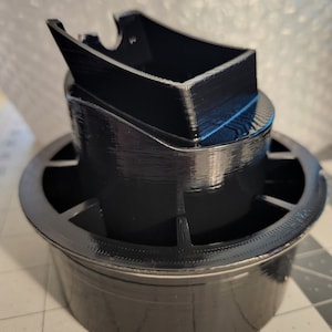 以下が含まれることがあります： 円形のベースと長方形のトップを持つ、黒色の3Dプリントされたオブジェクト。ベースはエッジの周りに一連のスロットがあります。トップはオープンで、エッジの周りにリップがあります。