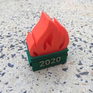 May include: A 3D printed ornament shaped like a trash can with a flame on top. The trash can is green and the flame is orange. The year 2020 is printed on the side of the trash can.