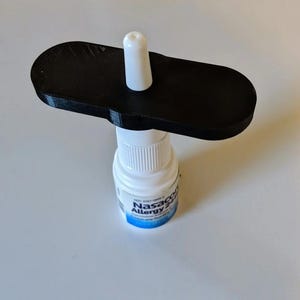 May include: A black 3D-printed device is placed on top of a white nasal spray bottle. The device has a flat, rectangular shape with rounded edges and a white cylindrical post. The bottle has blue text that reads "Nasacort Allergy 24HR".