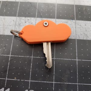 May include: Orange cloud-shaped key holder with a silver key and a metal ring. The key is inserted into the bottom of the cloud. A small metal rivet secures the key holder. The key holder is made of plastic.